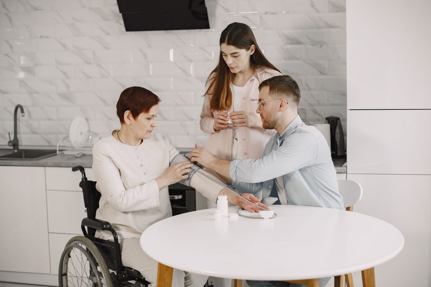 A caregiver and a nurse help a woman in a wheelchair check her blood pressure at home, with her medication on the kitchen table